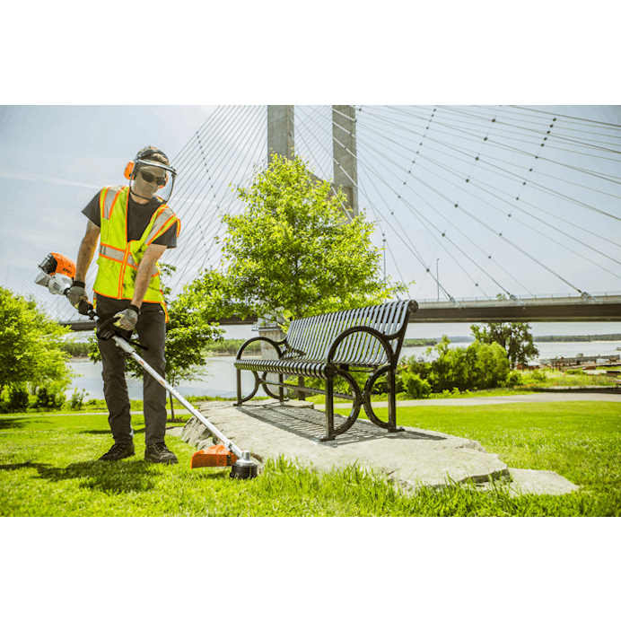 Man using the KM 91 R to trim grass near a bench in front of bridge 