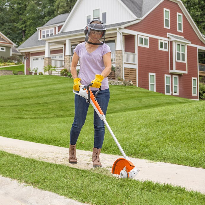Woman using STIHL FSA 30 to trim sidewalk grass wearing STIHL face guard